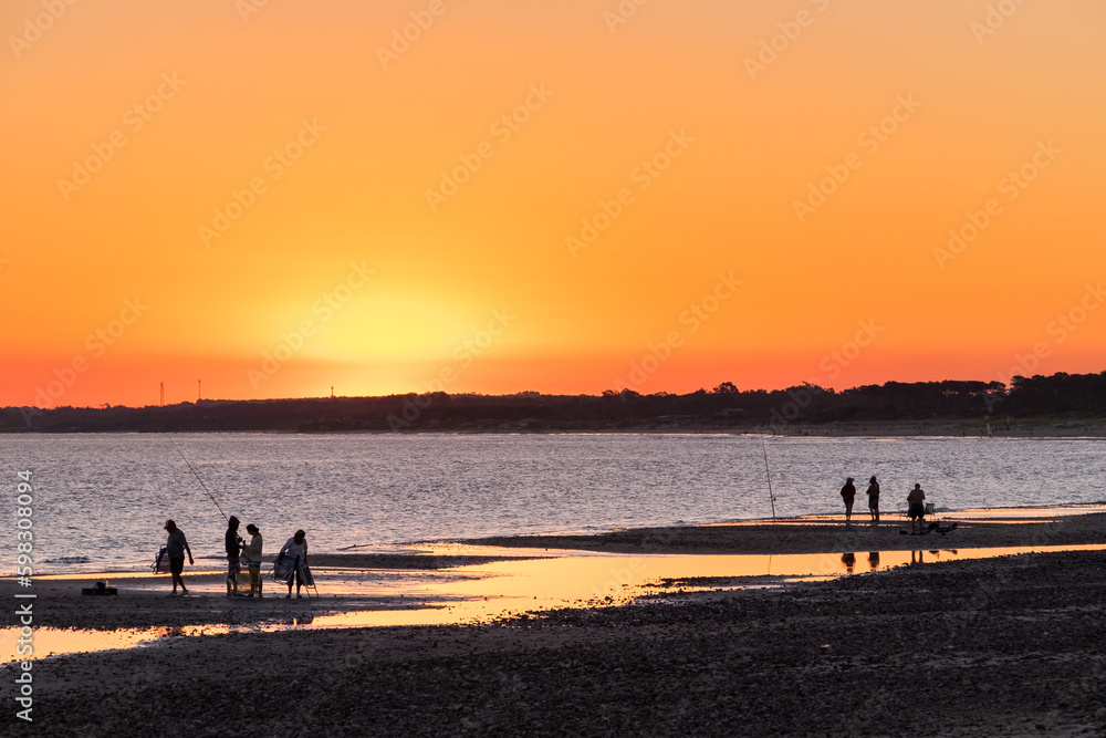 Silhouettes of fishermen on the beach of Las Flores, during a reddish sunset, with some puddles of water on the sand