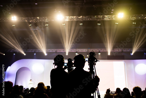Silhouette of the back of two cameramen standing shooting with video cameras to the stage where MC standing talking at the podium during the live event with people sitting watching the event in hall.