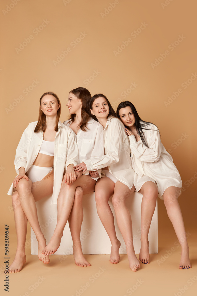 Naklejka premium Studio shot with group of young beautiful girls, women in white shirts sitting over beige color background. Healthy eating