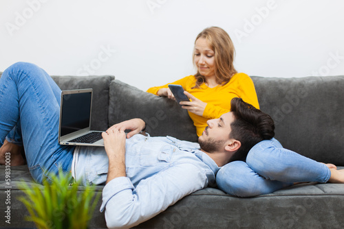 lovery couple at home on a sofa. boy lying with laptop on girlfriend's lap. girl looking at phone. photo