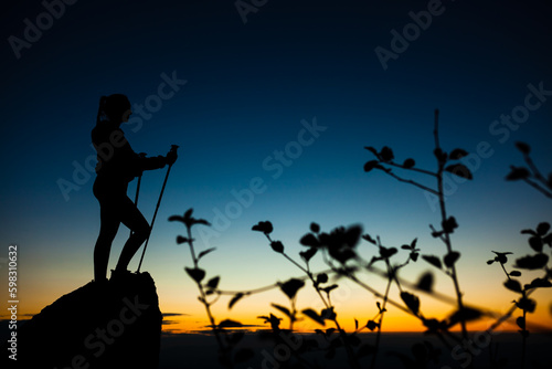 Silhouette of a hiker girl on a rock pedestal with hands up. Beautiful orange sunset on blue sky. Tree branches with leaves in the foreground.