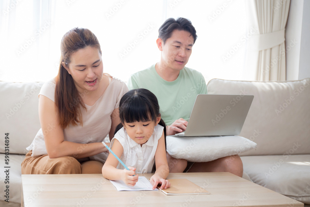 Happy family with mother teaching homework with daughter and father working with laptop on sofa in living room at home, mom explaining schoolwork with kid together, lifestyles and education.