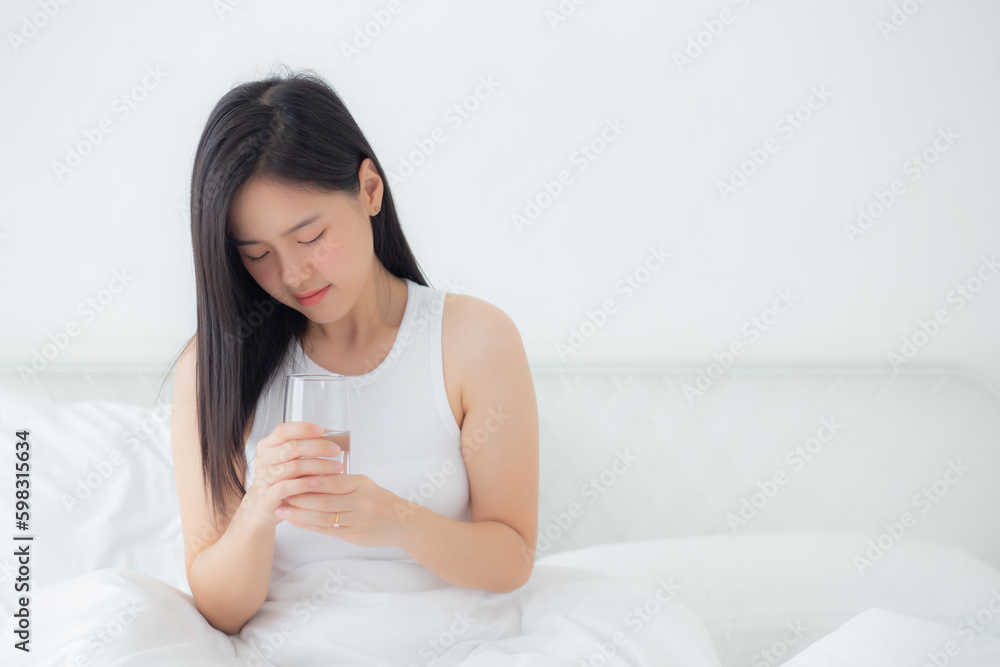 Beautiful young asian woman sitting on bed holding water of glass and drinking in the morning in bedroom at home, female drinking beverage mineral for healthy and nutrition, lifestyles concept. Beautiful young asian woman sitting on bed holding water of glass and drinking in the morning in bedroom at home, female drinking beverage mineral for healthy and nutrition, lifestyles concept.