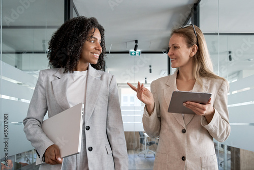  Two businesswoman working together, while walking in office and holding digital gadget pc. Small creative diversity team of African American and blond females executives meeting discuss work project. photo