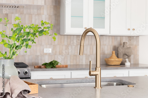 Canvastavla A kitchen faucet detail with a grey stone countertop island, gold faucet, stainless steel appliances, brown tile backsplash, and white cabinets