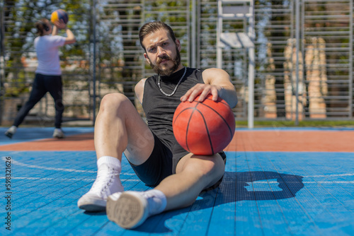 Portrait of tired athletic lebanese man in sportswear after playing basketball game. Guy sitting relaxing on urban city court. Fitness routine on sports field. Motivation. Outdoors at sunny morning © Andrii Iemelianenko