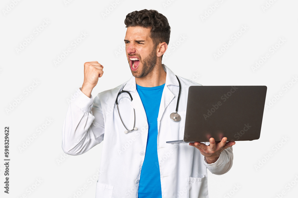 Young doctor man holding a laptop raising fist after a victory, winner concept. Young doctor man holding a laptop raising fist after a victory, winner concept.