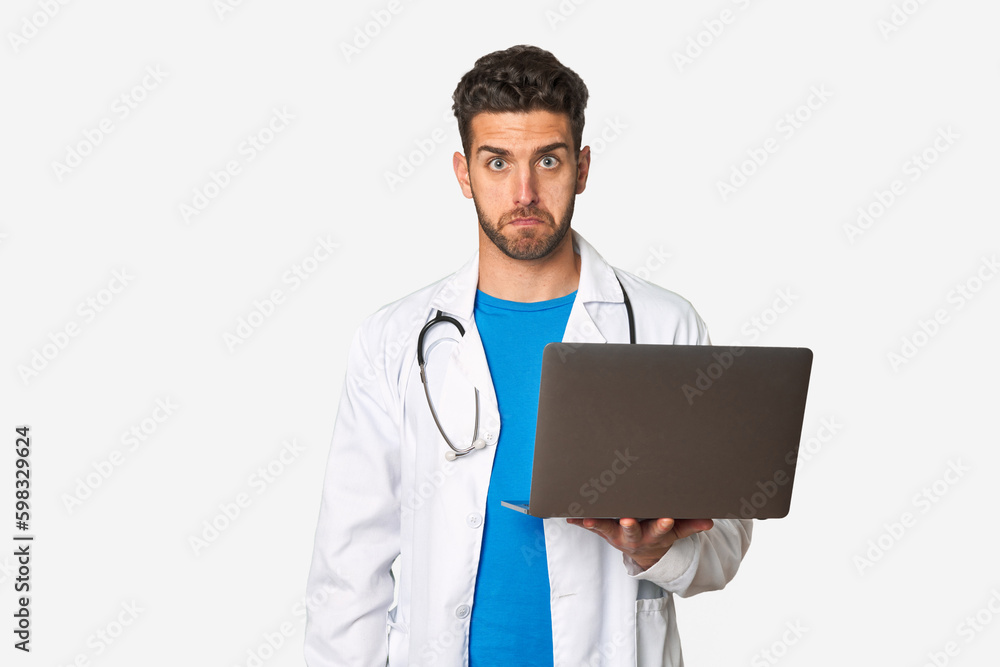 Young doctor man holding a laptop shrugs shoulders and open eyes confused. Young doctor man holding a laptop shrugs shoulders and open eyes confused.