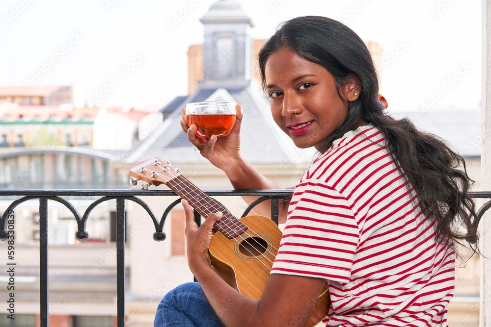 Indian woman playing ukulele on windowsill drinking a tea cup Indian woman playing ukulele on windowsill drinking a tea cup