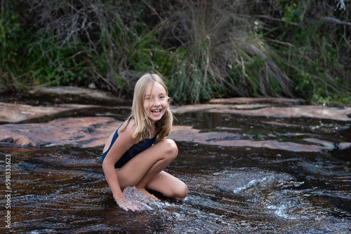 A happy girl in her swimming costume sitting in shallow river with water sliding over rocks during a family vacation