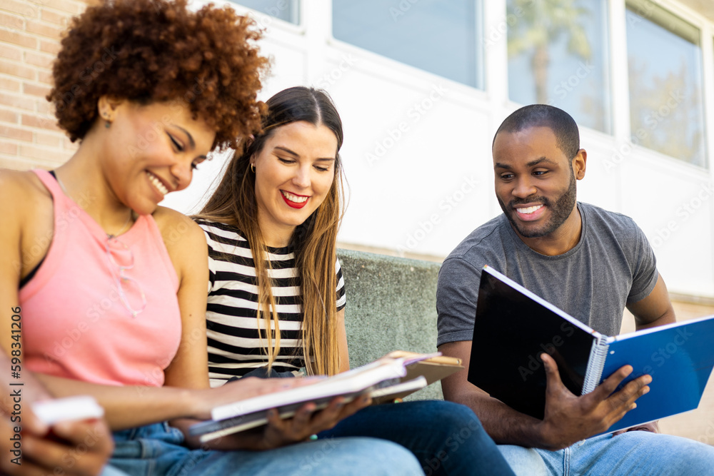 Fototapeta premium A group of young multi-ethnic students outside the university.The 3 young people sit happily looking at the book of the afro-haired girl to study for an exam.Concept of multiracial university students