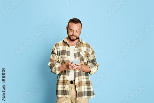 Portrait of happy, smiling, bearded man in checkered shirt typing messages with mobile phone against blue studio background. Concept of human emotions, lifestyle, facial expression. Ad