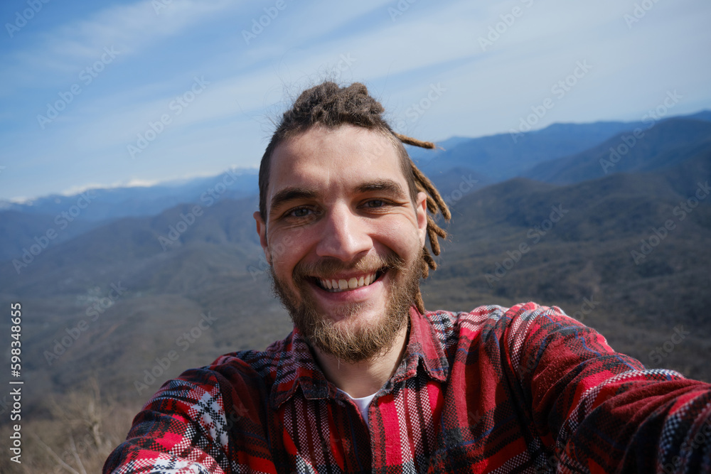 Concept of healthy active lifestyle. Young travel blogger with dreadlocks went hiking in mountains. Man enjoys views of nature takes video or selfie photo on phone or camera. Wide-angle view portrait.