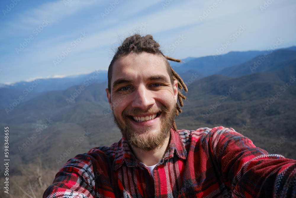 Concept of healthy active lifestyle. Young travel blogger with dreadlocks went hiking in mountains. Man enjoys views of nature takes video or selfie photo on phone or camera. Wide-angle view portrait. Concept of healthy active lifestyle. Young travel blogger with dreadlocks went hiking in mountains. Man enjoys views of nature takes video or selfie photo on phone or camera. Wide-angle view portrait.