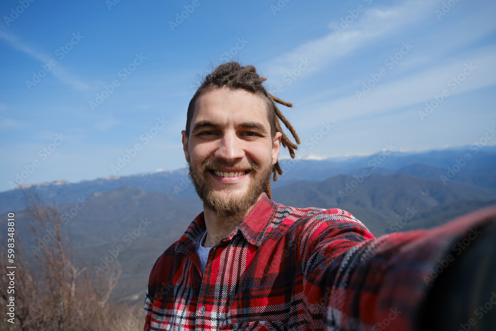 Concept of healthy active lifestyle. Young travel blogger with dreadlocks went hiking in mountains. Man enjoys views of nature takes video or selfie photo on phone or camera. Wide-angle view portrait. Concept of healthy active lifestyle. Young travel blogger with dreadlocks went hiking in mountains. Man enjoys views of nature takes video or selfie photo on phone or camera. Wide-angle view portrait.