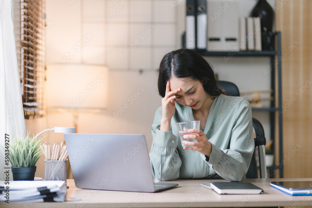 The stressed and exhausted millennial Asian businesswoman is seen sitting at her office desk with her hand on her head, indicating a hard working day where she is overloaded with work. The stressed and exhausted millennial Asian businesswoman is seen sitting at her office desk with her hand on her head, indicating a hard working day where she is overloaded with work.