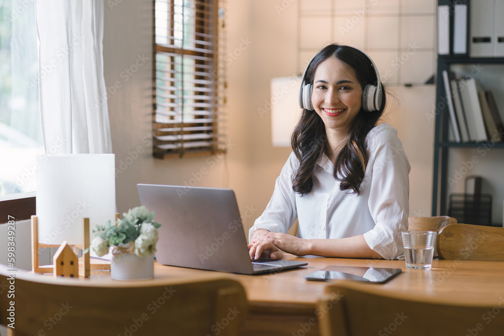 Image of a young, pleased, happy, cheerful, cute, and beautiful businesswoman sitting indoors at a dining table in her home office, using a laptop computer, and listening to music with earphones. Image of a young, pleased, happy, cheerful, cute, and beautiful businesswoman sitting indoors at a dining table in her home office, using a laptop computer, and listening to music with earphones.