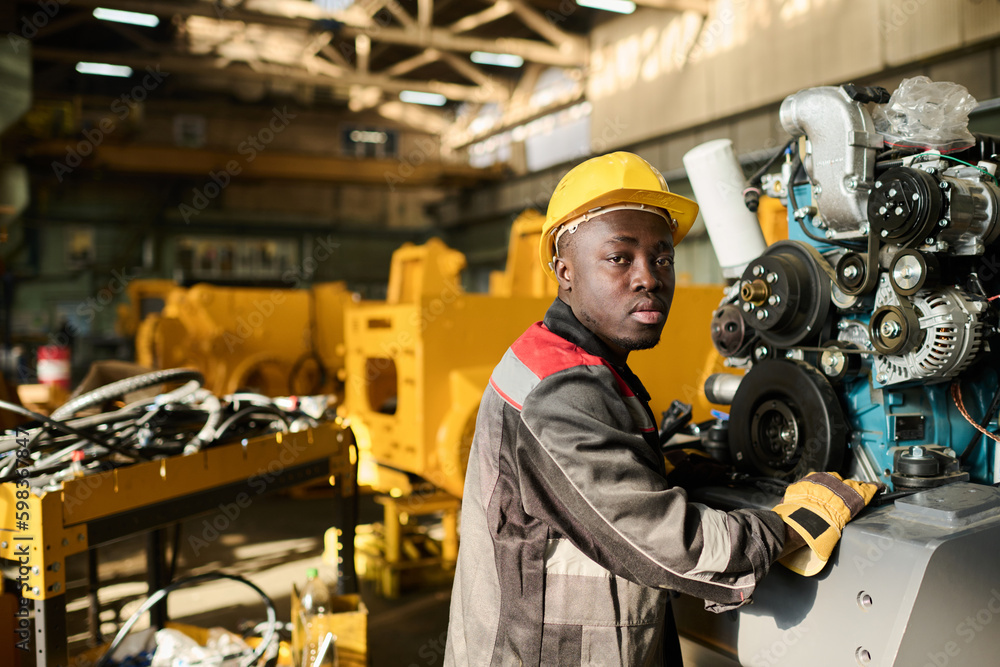 Young African American engineer in workwear and yellow protective helmet looking at camera while operating huge industrial machine Young African American engineer in workwear and yellow protective helmet looking at camera while operating huge industrial machine
