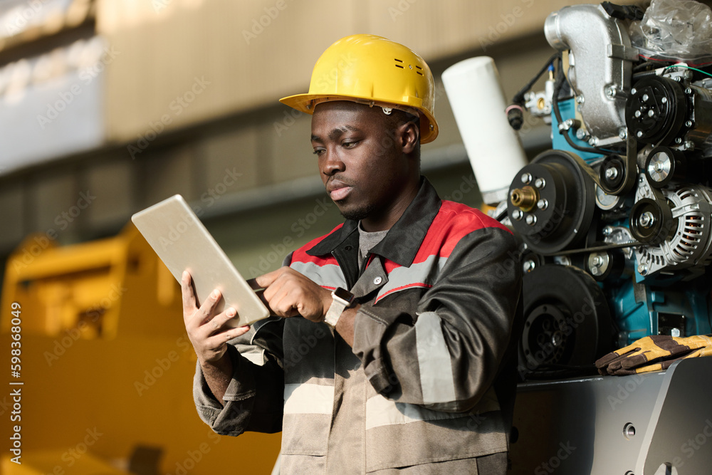 Serious young foreman or control manager in safety helmet and workwear scrolling through online technical information in tablet Serious young foreman or control manager in safety helmet and workwear scrolling through online technical information in tablet