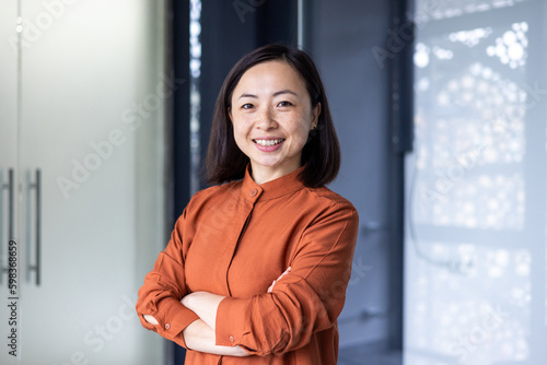 Portrait of young beautiful and successful Asian business woman, female employee smiling and looking at camera with crossed arms, financial woman satisfied with the results of her work achievement.