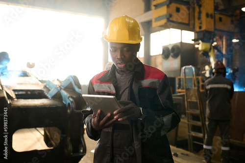 African American engineer or foeman scrolling in tablet while standing in front of camera in workshop and looking through online manual