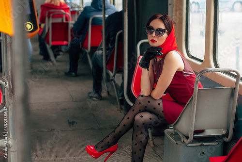 A stylish woman in sunglasses, in a red dress, black leather gloves, tights, red shoes and a scarf, sits in a tram and looks out the window.