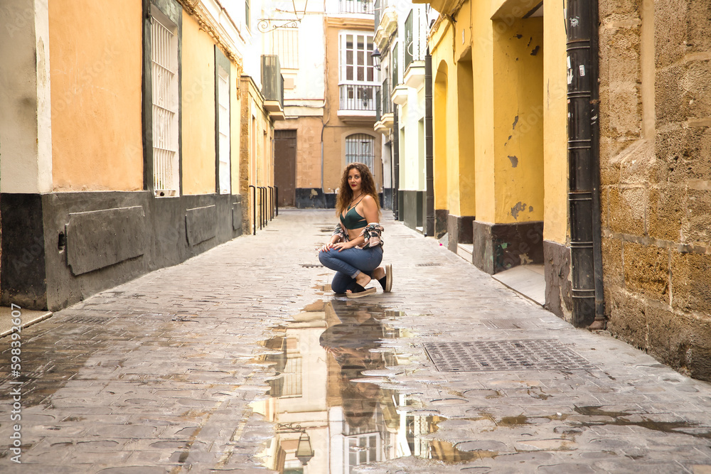 Young and beautiful woman, blonde, with curly hair and blue eyes, wearing floral shirt, green top and jeans, crouched sensually posing on a lonely street, reflected in a puddle.