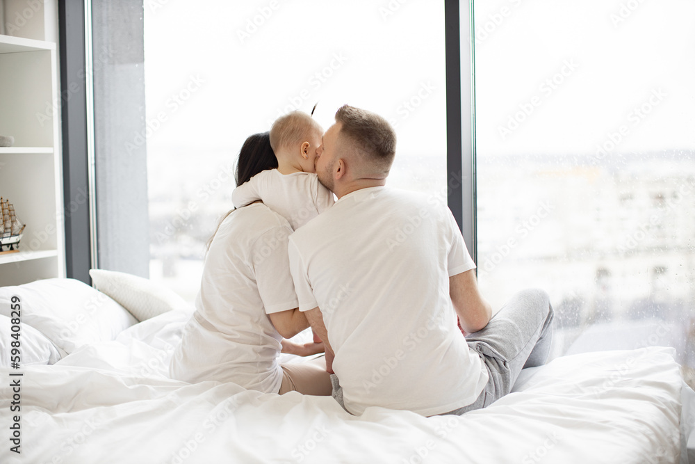 Rear view of loving father kissing his baby daughter while little girl hugging caring mom in morning after waking up. Young married couple with kid resting on white blanket in bedroom of modern flat. Rear view of loving father kissing his baby daughter while little girl hugging caring mom in morning after waking up. Young married couple with kid resting on white blanket in bedroom of modern flat.