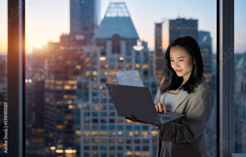 Young happy busy Asian business woman executive working on laptop at night in dark corporate office. Professional businesswoman manager using computer standing at window with big city Generative AI