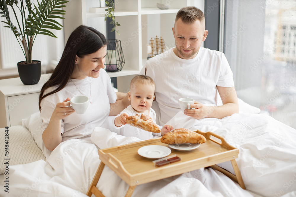Happy husband and wife holding coffee cups while cute kid reaching out for delicious food on bed tray in modern studio flat. Small caucasian family having enjoyable meal together in home interior. Happy husband and wife holding coffee cups while cute kid reaching out for delicious food on bed tray in modern studio flat. Small caucasian family having enjoyable meal together in home interior.
