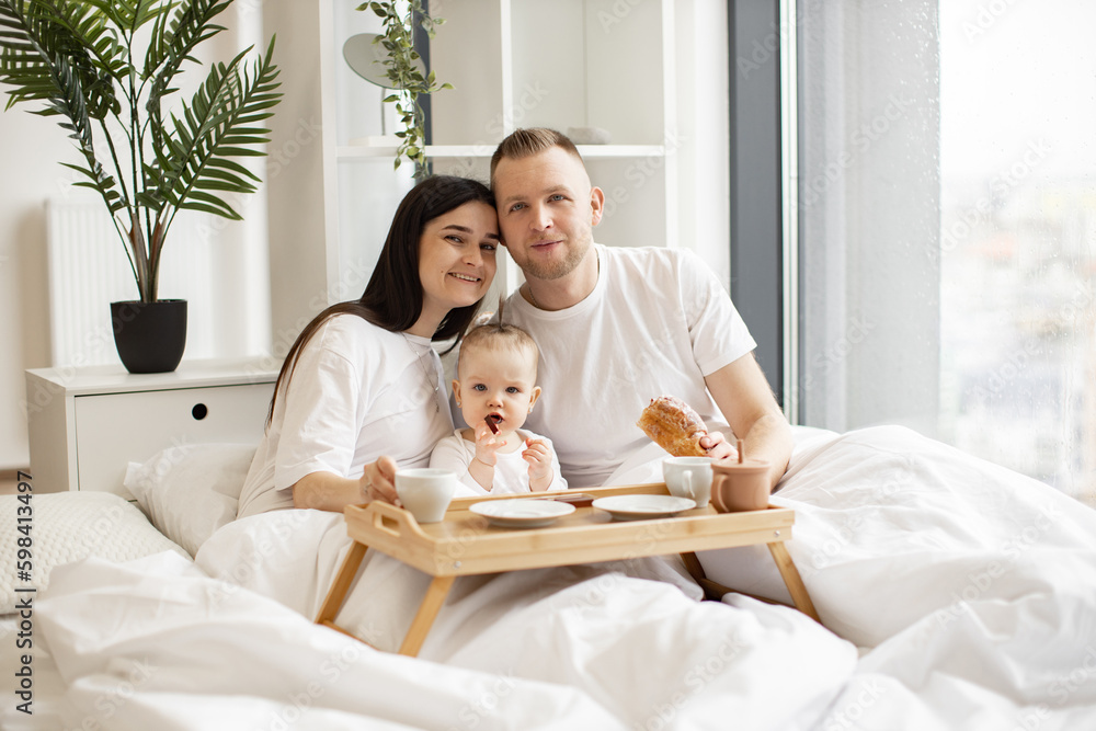 Smiling parents posing with breakfast on bed tray while sweet baby girl eating healthy snack and staying under soft blanket in bright room. Loving married couple with baby eating early meal indoors. Smiling parents posing with breakfast on bed tray while sweet baby girl eating healthy snack and staying under soft blanket in bright room. Loving married couple with baby eating early meal indoors.