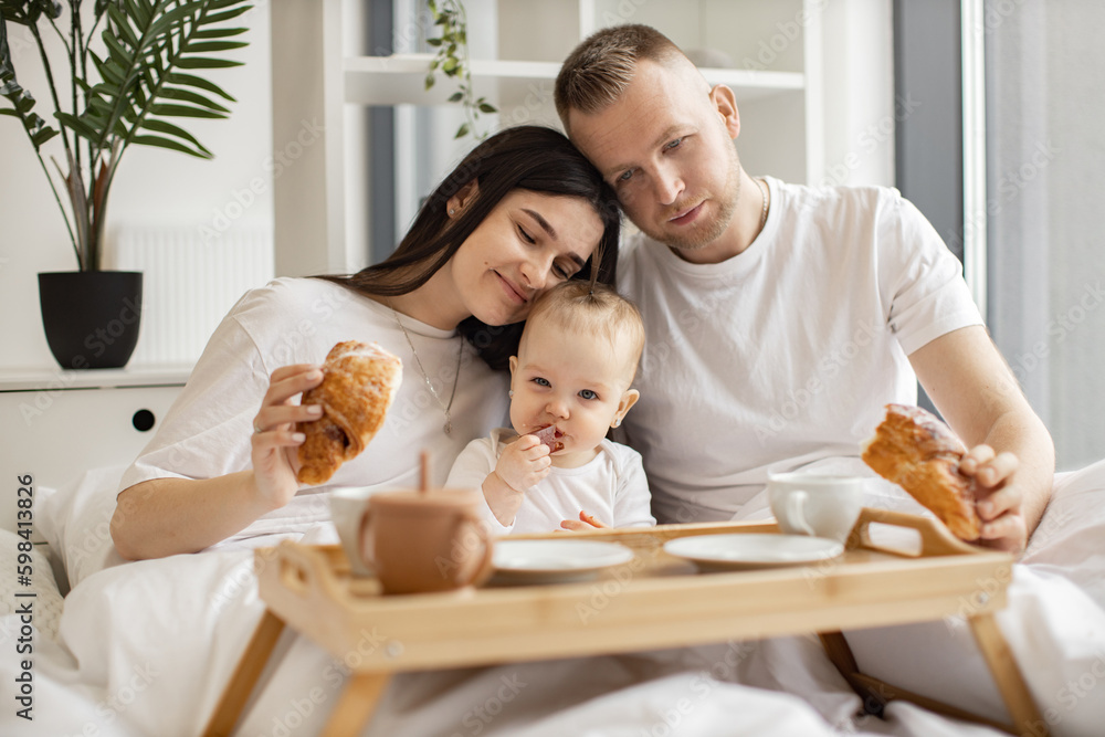 Loving spouses in everyday wear holding croissants while cuddling little daughter that eating fruit candy in bedroom interior. Charming family of three sharing closeness during weekend rest. Loving spouses in everyday wear holding croissants while cuddling little daughter that eating fruit candy in bedroom interior. Charming family of three sharing closeness during weekend rest.