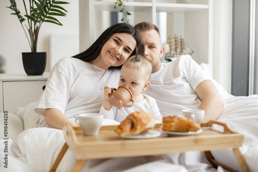 Sweet little baby drinking from sippy cup while joyful brunette woman snuggling with blonde male in cozy bedroom with tray on foreground. Caring family spending quality time together at home. Sweet little baby drinking from sippy cup while joyful brunette woman snuggling with blonde male in cozy bedroom with tray on foreground. Caring family spending quality time together at home.