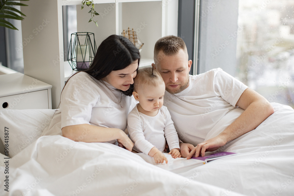 Young married couple in casual outfit sitting in cozy bed with little daughter and reading interactive baby book on Sunday morning. Loving parents making story come alive for curious daughter.