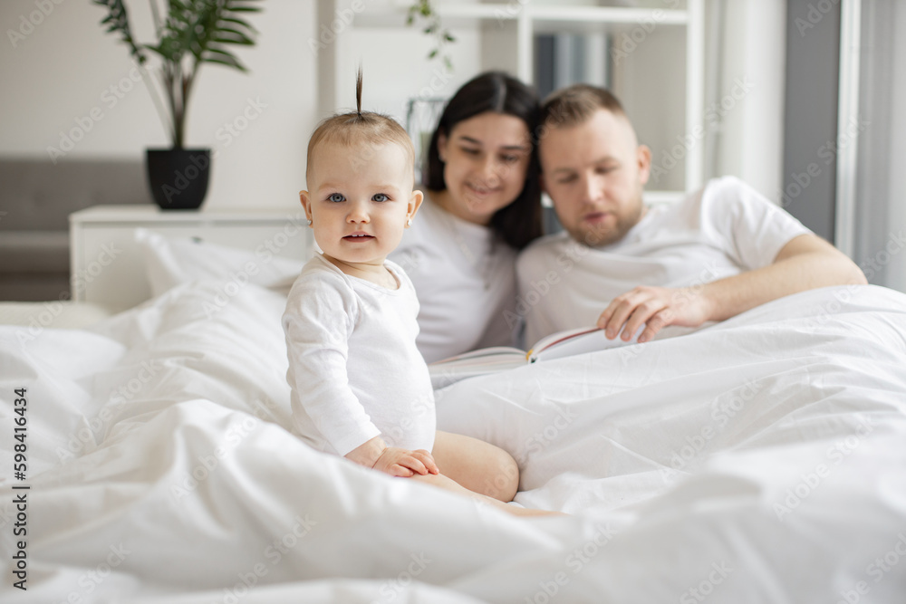 Cheerful little kid in white baby clothes resting on bed while joyful adult people staying under blanket in bright bedroom interior. Loving mother and father telling nursery rhyme from verse book. Cheerful little kid in white baby clothes resting on bed while joyful adult people staying under blanket in bright bedroom interior. Loving mother and father telling nursery rhyme from verse book.