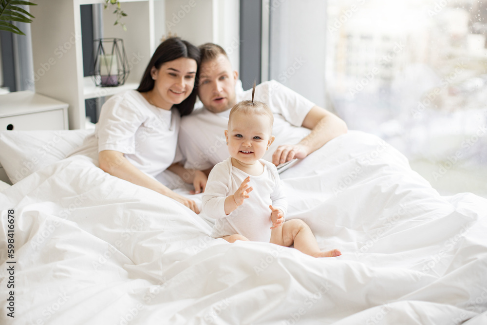 Cheerful little kid in white baby clothes resting on bed while joyful adult people staying under blanket in bright bedroom interior. Loving mother and father telling nursery rhyme from verse book.
