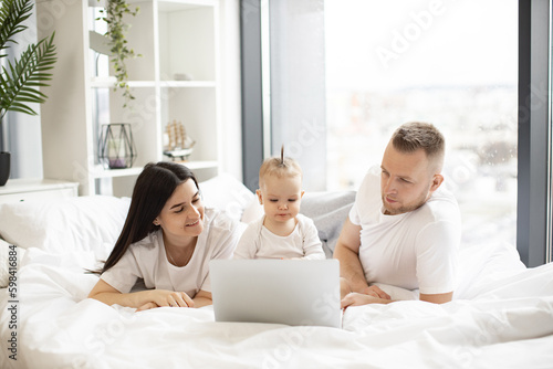 Young caucasian parents in everyday wear lying on bed with cute infant daughter in front of portable computer in home interior. Affectionate family of three watching movie via internet on sunny day.