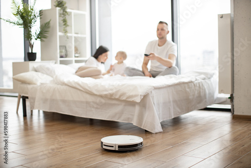 Brunette woman playing with infant girl on bed while young man turning on robot vacuum cleaner via remote control in studio room. Modern family resting at weekend using one touch home cleaning.