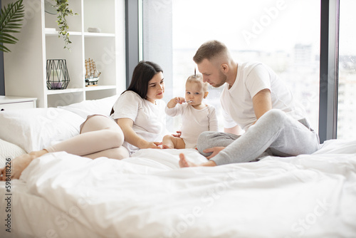 Charming baby girl eating with spoon by herself while sitting on bed with mother and father in casual attire on sunny day. Attentive parents taking care of little daughter while staying at home.