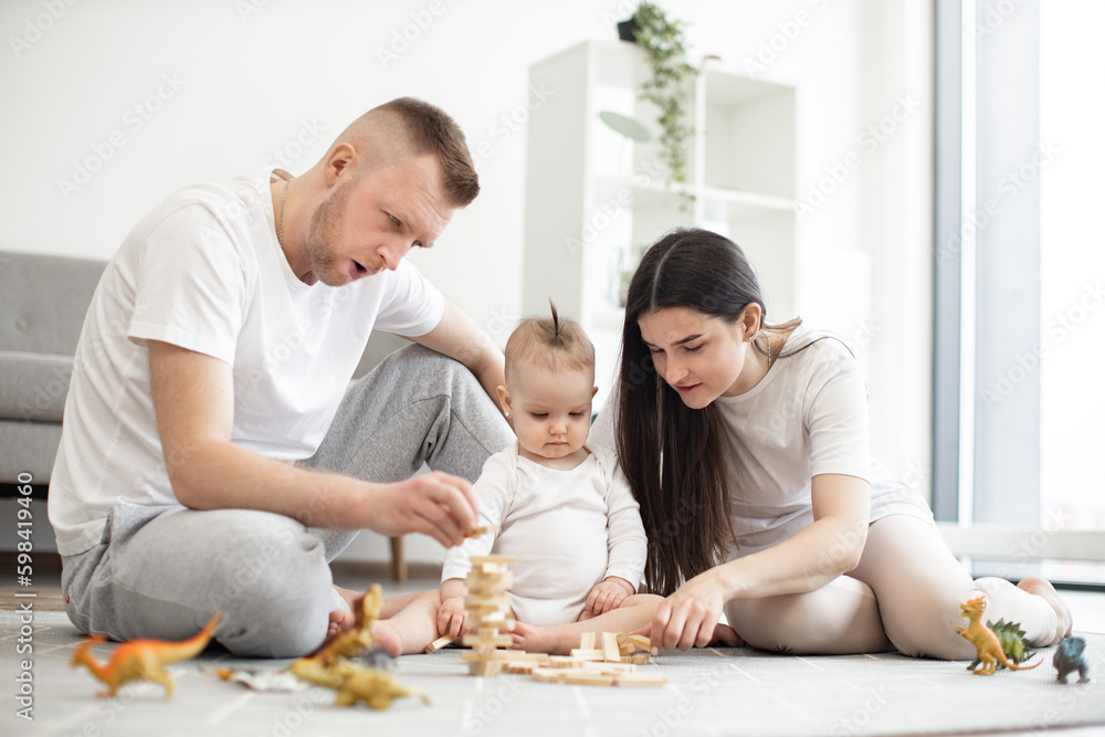 Loving caucasian parents in leisurewear sitting with small kid in white bodysuit and building toy tower from wooden blocks on floor of living room. Excited family bonding together using board game.