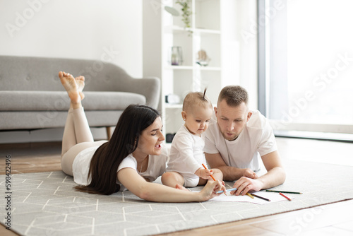 Adorable little baby girl in white cozy bodysuit holding red pencil while sitting on floor with mindful mother and father on both sides. Adult people and kid enjoying first scribbles on paper at home.