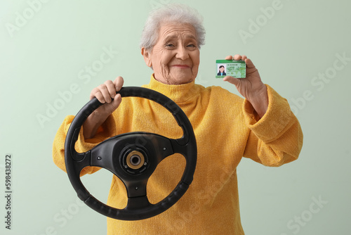 Senior woman with steering wheel and driving license on green background