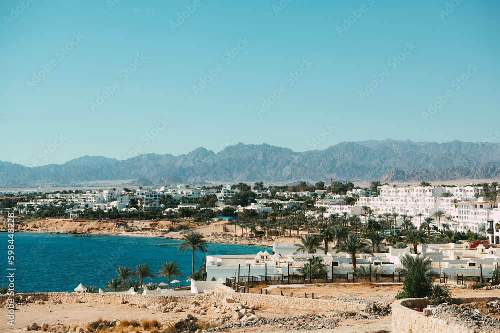 An enchanting view unfolds as azure waters meet endless desert sands, adorned by pristine white buildings and swaying palm trees, a picturesque oasis.