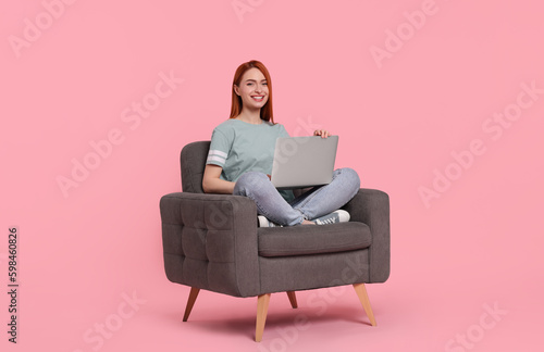 Smiling young woman with laptop sitting in armchair on pink background