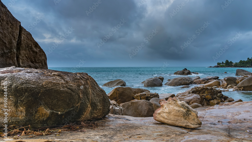 Picturesque granite boulders are scattered on the beach. Turquoise ocean waves foam around the stones. A green hill on the horizon. Clouds in the sky. Seychelles. Mahe.