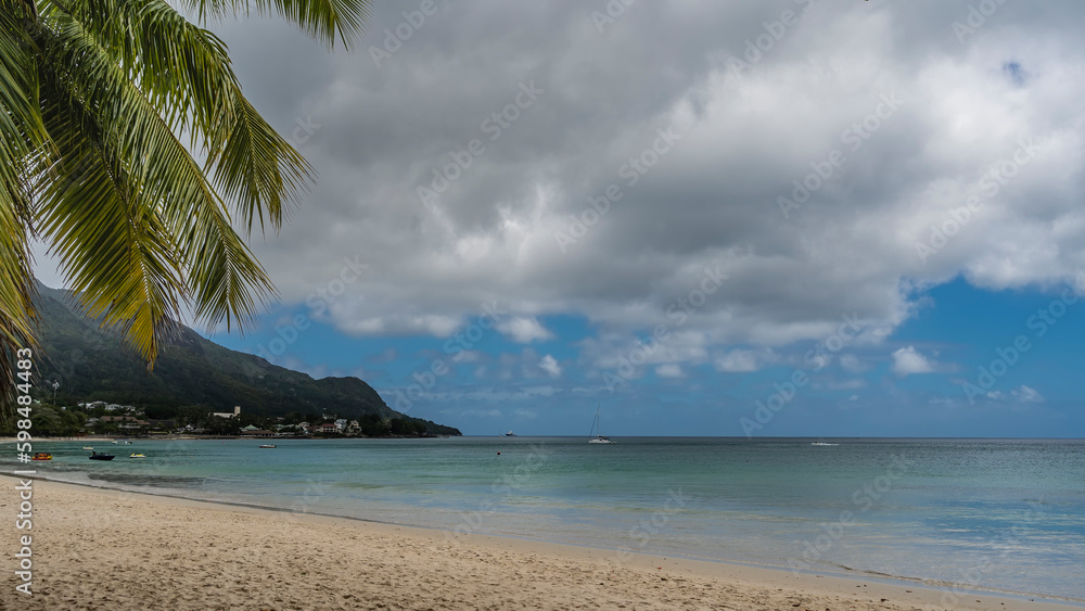 Yachts and boats are visible in the calm turquoise ocean. A green hill in the distance. Footprints on a sandy beach. Lush palm leaves against the sky and clouds. Seychelles. Mahe. Beau Vallon