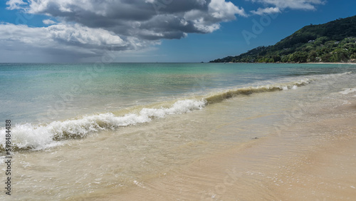 The wave rolls towards the shore  swirling with spray and foam. The water mixes with the sand of the beach. The turquoise ocean is calm. A green hill against the blue sky and clouds. Seychelles.