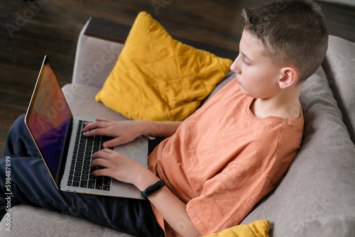 Teen boy study or play game on laptop home interior background. Guy doing homework and typing on laptop