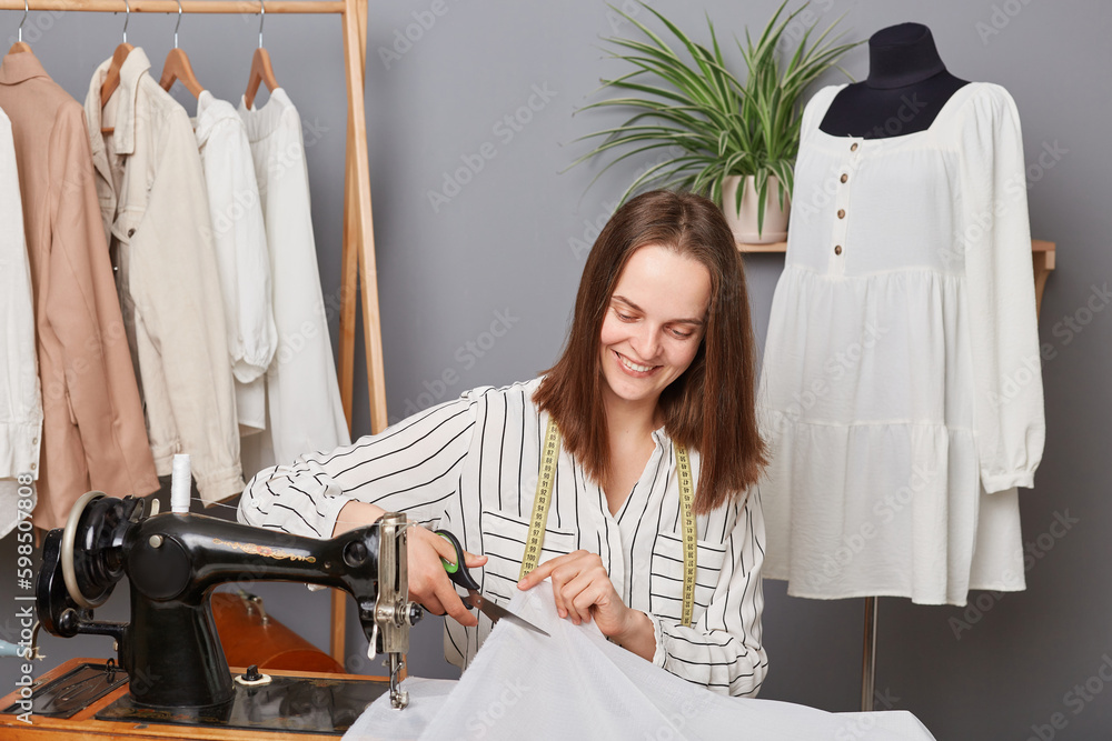 Attractive joyful brown haired woman seamstress cutting textile sewing dress or shirt, process of creation trendy clothing, smiling happily enjoying her job. Attractive joyful brown haired woman seamstress cutting textile sewing dress or shirt, process of creation trendy clothing, smiling happily enjoying her job.