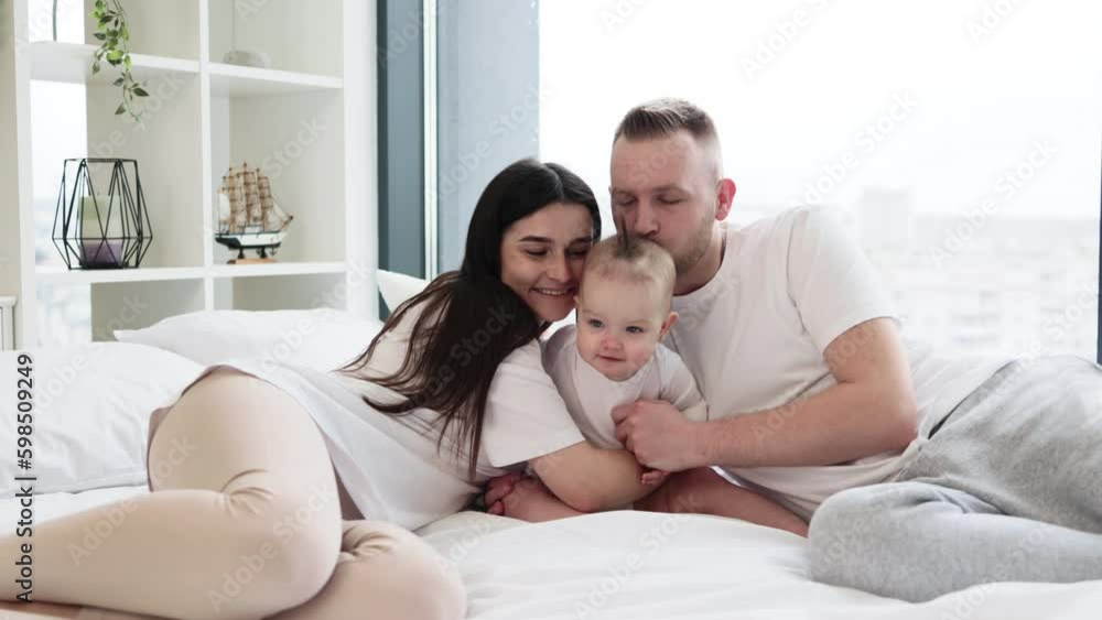 Young parents resting with pretty little daughter on comfy bed with panoramic windows on background. Loving mother and father embracing their toddler from both sides and smiling on camera. Young parents resting with pretty little daughter on comfy bed with panoramic windows on background. Loving mother and father embracing their toddler from both sides and smiling on camera.